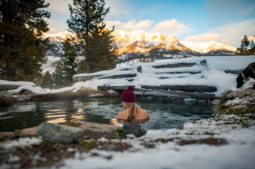 A person in a beanie relaxes in a steaming hot spring surrounded by snow-covered mountains and trees.