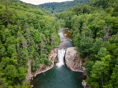 Aerial view of a waterfall cascading into a serene river, surrounded by lush green trees and mountains.