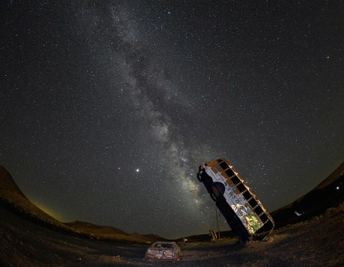 A starry night sky with the Milky Way, featuring two abandoned cars in the foreground.