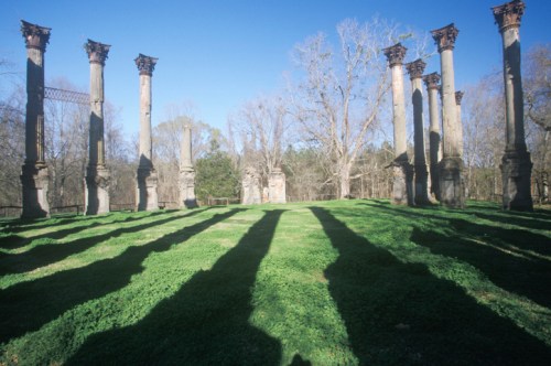 Ruins of tall columns casting long shadows on green grass under a clear blue sky.