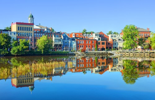 Colorful buildings reflect in calm water under a clear blue sky, creating a picturesque waterfront scene.