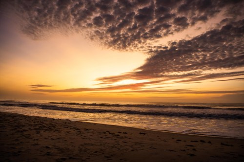 A serene beach at sunset, with vibrant clouds reflecting orange and purple hues over gentle waves.
