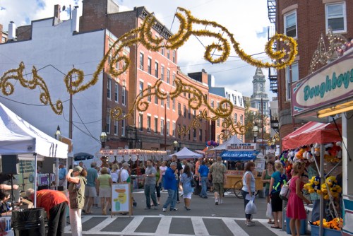 A bustling street fair with colorful stalls, decorations overhead, and crowds enjoying food and festivities.