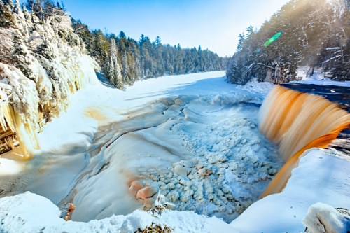 A winter landscape featuring a frozen waterfall cascading into a snowy river, surrounded by trees and bright blue skies.