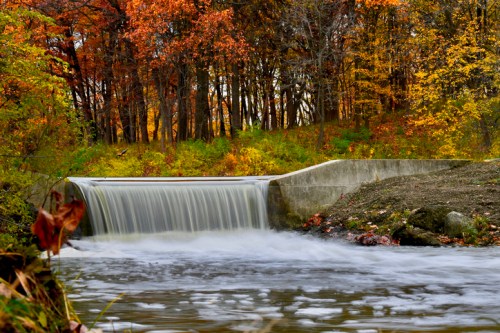 A serene waterfall flows over a concrete barrier, surrounded by vibrant autumn foliage and a calm river.