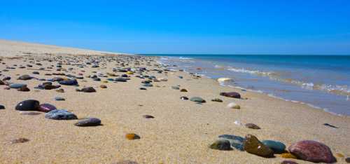 A sandy beach scattered with colorful pebbles, with gentle waves lapping at the shore under a clear blue sky.