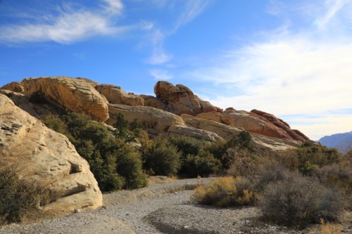 A winding path leads through rocky terrain and shrubs under a blue sky with wispy clouds.