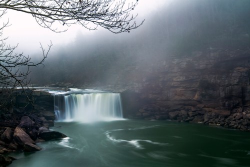 A misty waterfall cascades into a green pool, surrounded by rocky cliffs and bare trees.