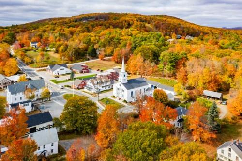Aerial view of a small town with colorful autumn foliage, featuring a white church and quaint buildings.