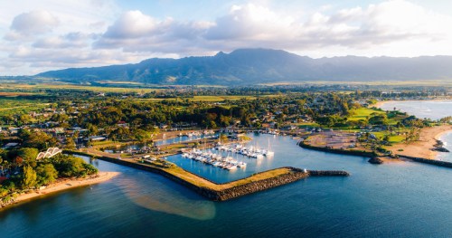 Aerial view of a marina with boats, surrounded by lush greenery and mountains under a partly cloudy sky.