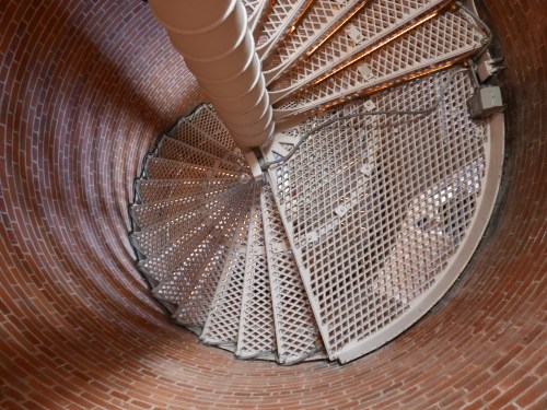 A spiral staircase viewed from above, featuring a metal grate and brick walls surrounding it.