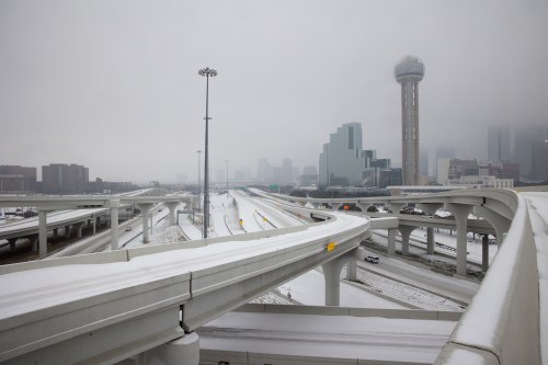 Snow-covered highways and overpasses with a city skyline in the background, partially obscured by fog.