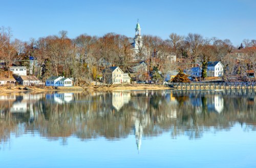 A serene lakeside view with trees and houses reflecting on calm water, featuring a church steeple in the background.