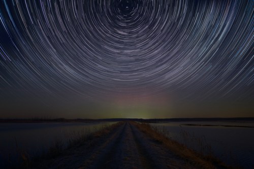 A starry night sky with circular star trails above a calm lake and a dirt road leading into the distance.