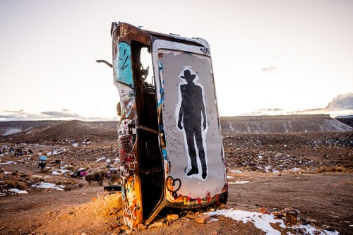 A graffiti-covered van door stands in a desert landscape, featuring a silhouette of a person against a sunset sky.