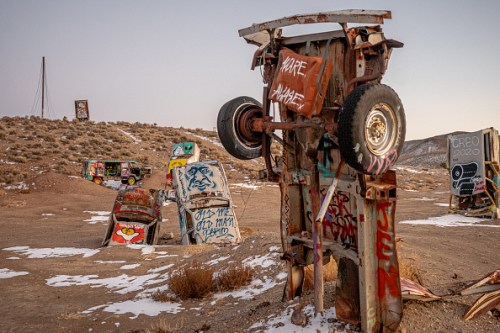 A collection of rusted, graffiti-covered cars in a desert landscape, some overturned, with sparse vegetation and distant hills.
