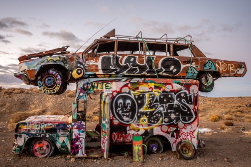 A colorful, graffiti-covered car stacked on top of an old van in a desert landscape.