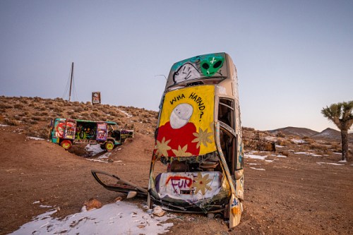A colorful, graffiti-covered abandoned bus in a desert landscape, with another painted vehicle in the background.