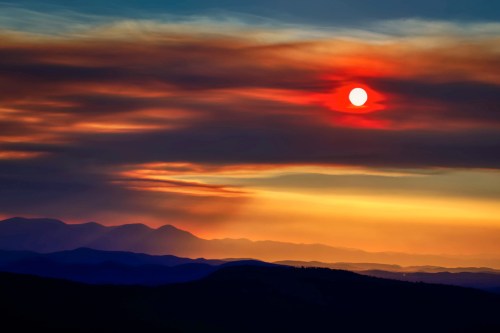 A vibrant sunset with a red sun setting behind layered mountains and colorful clouds in the sky.