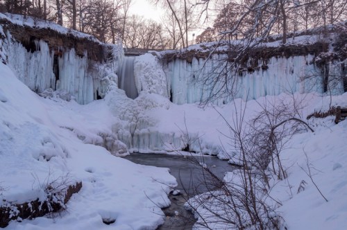 A frozen waterfall cascading over icy cliffs, surrounded by snow-covered ground and bare trees.