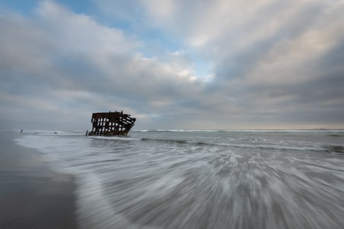 A shipwreck partially submerged in a beach, with waves gently washing over the sand under a cloudy sky.