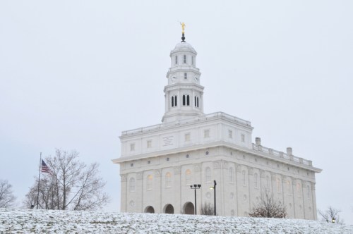 A white historic building with a golden statue on top, surrounded by snow and trees, under a gray sky.
