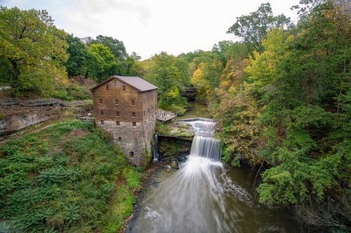A historic wooden mill beside a flowing waterfall, surrounded by lush green trees and autumn foliage.