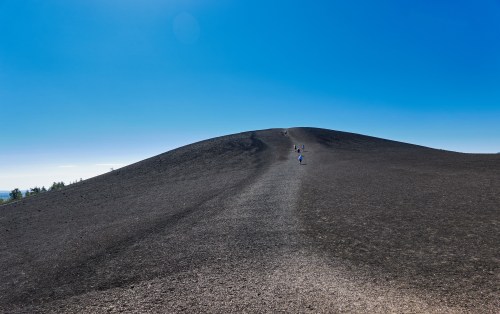 A wide view of a volcanic hill with a clear blue sky, featuring hikers ascending the dark, textured slope.