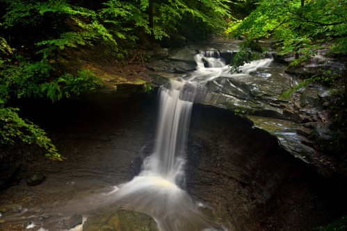 A serene waterfall cascades over rocky ledges, surrounded by lush green foliage in a tranquil forest setting.