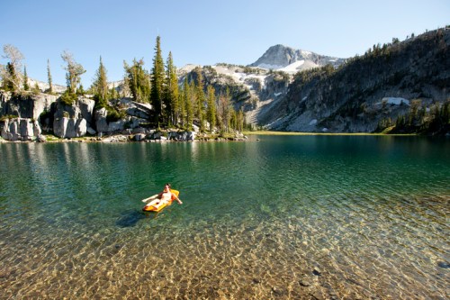 A person kayaking on a clear lake surrounded by mountains and trees under a bright blue sky.