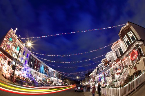 Colorful holiday lights adorn houses along a street, with a dark blue sky and light trails from passing cars.