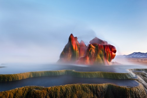Colorful rock formations rise from a misty landscape, surrounded by calm water and distant mountains under a clear sky.