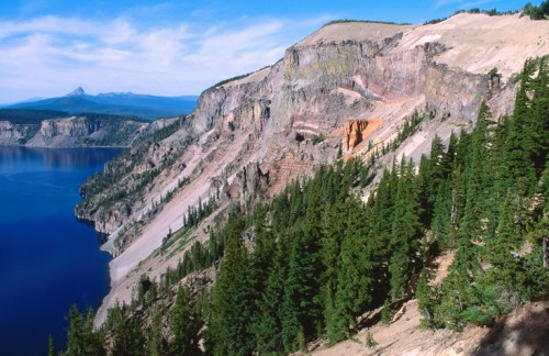 A scenic view of a steep cliffside overlooking a deep blue lake, surrounded by lush green trees and distant mountains.