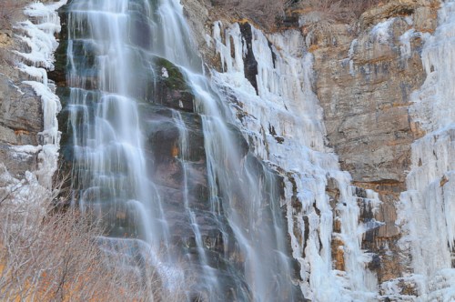 A cascading waterfall with icy formations on rocky cliffs, surrounded by bare trees.