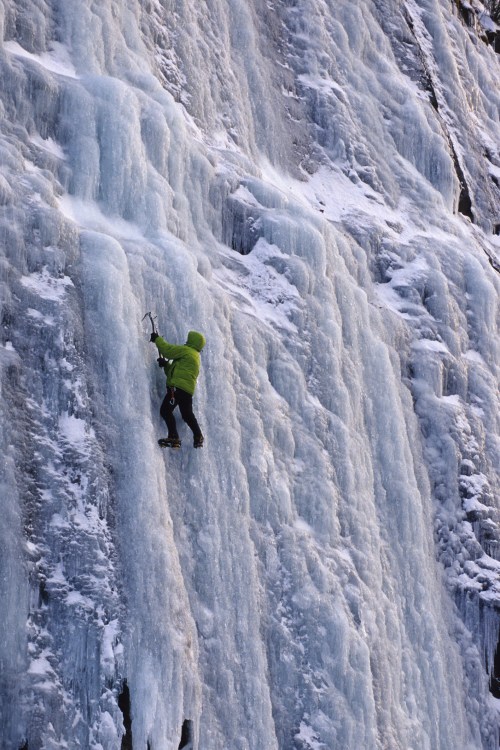 A climber in a green jacket ascends a frozen waterfall, surrounded by icy blue formations and snow.