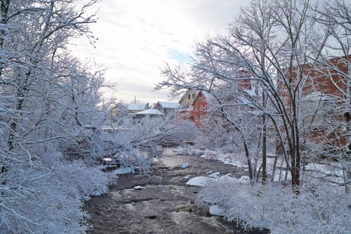 A serene winter scene with a snow-covered riverbank, frosted trees, and quaint buildings in the background.