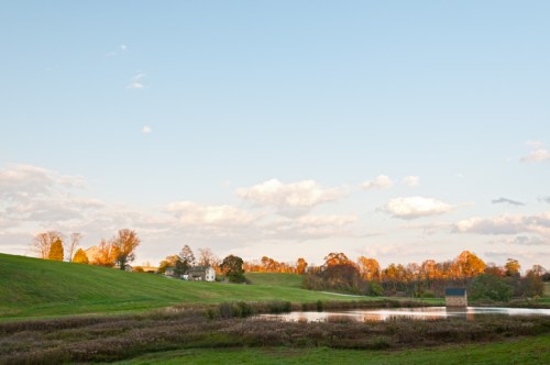 A serene landscape featuring a calm pond, green hills, and trees with autumn foliage under a clear blue sky.