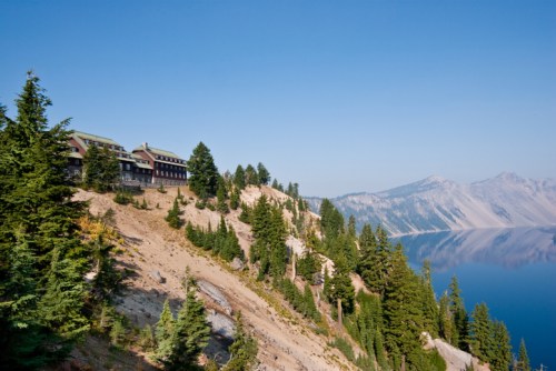 A scenic view of a lodge on a cliffside, surrounded by trees and overlooking a calm lake and mountains in the distance.