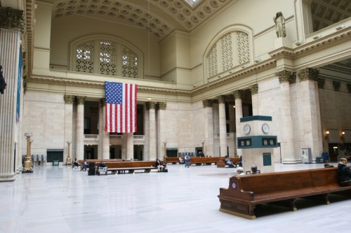 Interior of a grand train station featuring high ceilings, large columns, an American flag, and wooden benches.
