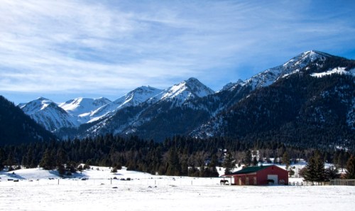 A snowy landscape with a red barn in the foreground and majestic mountains in the background under a blue sky.