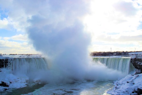 A powerful waterfall cascades over cliffs, creating a misty spray against a bright sky, with snow on the ground.