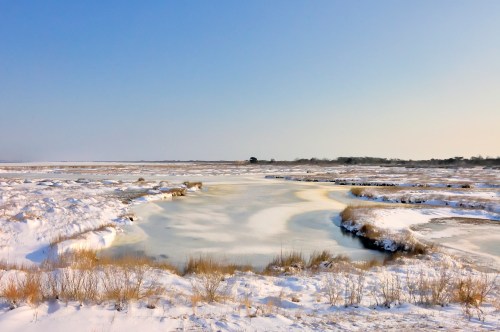 A serene winter landscape featuring a frozen river surrounded by snow-covered fields and a clear blue sky.
