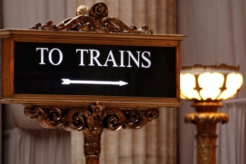 Ornate sign directing to trains, featuring an arrow pointing right, with a decorative lamp in the background.