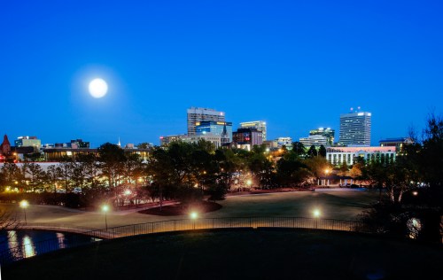 City skyline at dusk with a full moon, featuring illuminated buildings and a park in the foreground.