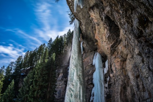A frozen waterfall cascading down a rocky cliff, surrounded by evergreen trees and a blue sky.