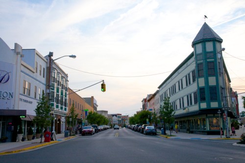 A street view featuring historic buildings, parked cars, and a cloudy sky in a small town setting.