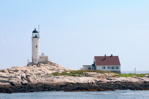 A lighthouse stands on rocky land beside a small house, with a clear blue sky in the background.