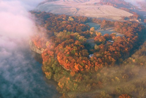 Aerial view of a colorful forested island surrounded by misty water, showcasing vibrant autumn foliage.