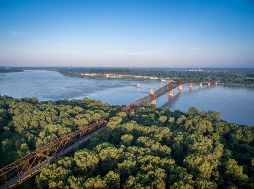 Aerial view of a rusted bridge spanning a river, surrounded by lush green trees and a clear blue sky.
