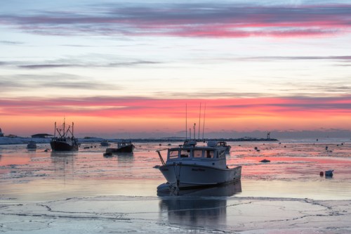 A serene sunset over a frozen harbor, with fishing boats silhouetted against vibrant pink and orange skies.